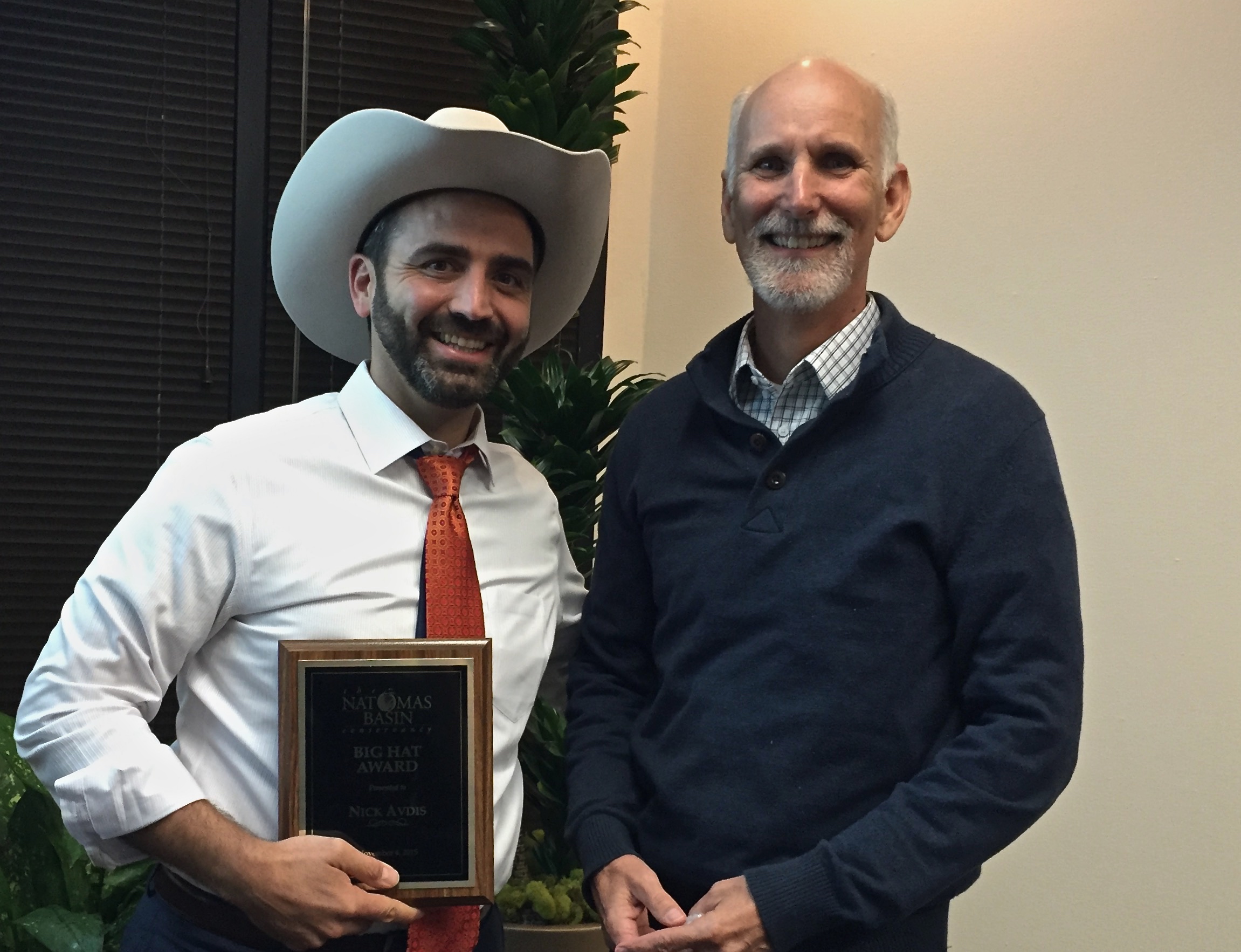two men standing and facing the camera, smiling, with the one on the left wearing a stetson hat and holding a wooden plaque award