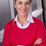 portrait of Michele McCormick standing with her arms folded, wearing a collard shirt with a red sweater over it and smiling directly at the camera