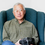 Gerry Tsurada sits on a blue high-backed chair wearing a green checkered button-down shirt and holding a camera in his lap