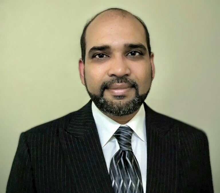 A photo of Chandra Chilmakuri wearing a black suit, white collared shirt and gray and black striped tie against a green yellow background.